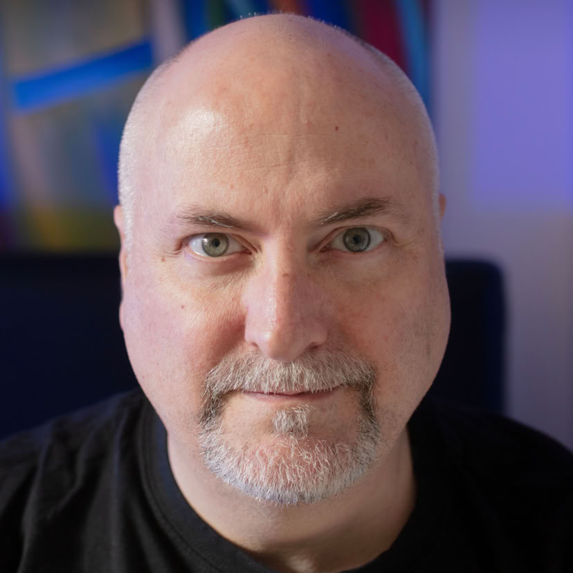 A bald man with a goatee, wearing a black shirt, is looking directly at the camera with a neutral expression. The background is blurred with abstract colors.